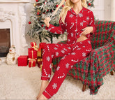 Woman in red Christmas pajamas sitting in a festive living room with a tree and fireplace.