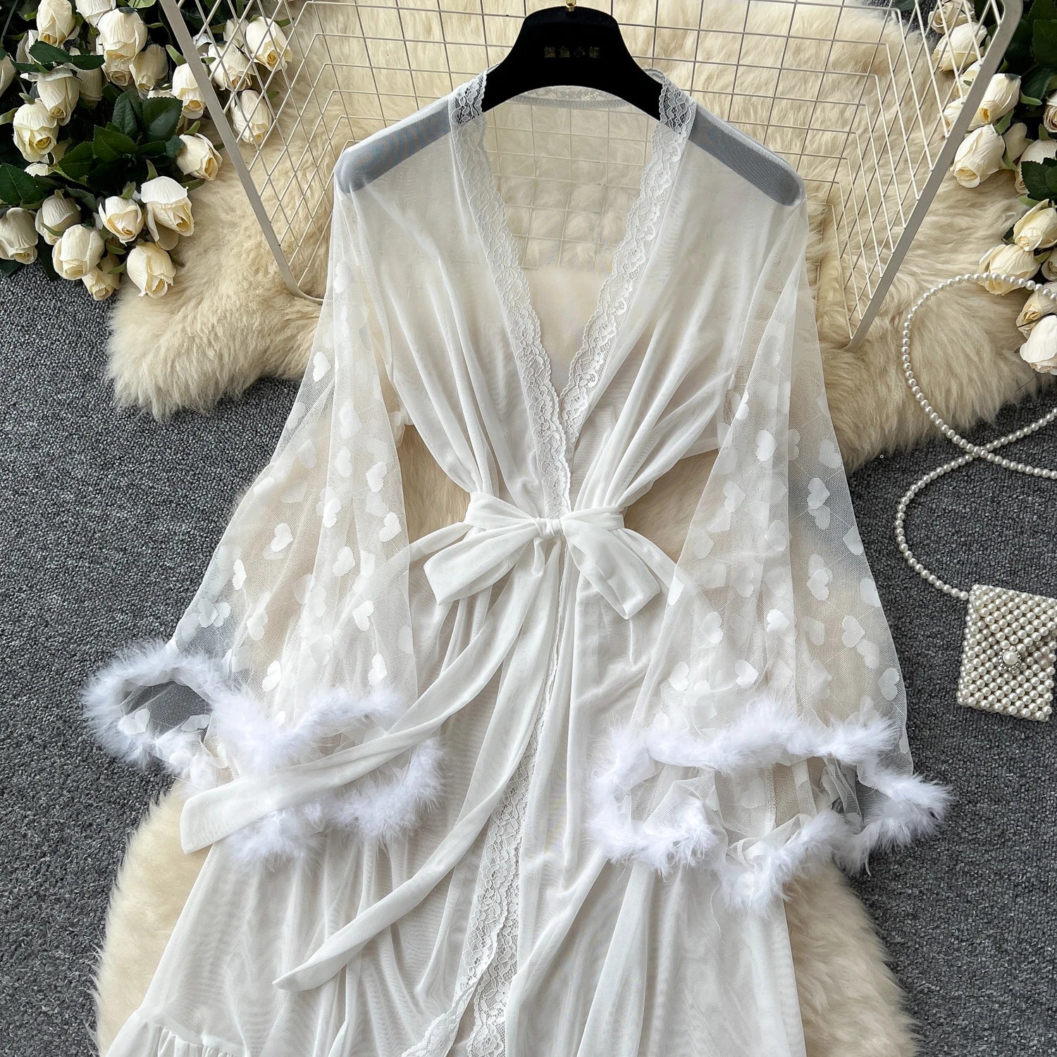 White lace robe with feather details on a hanger against a decorative background.