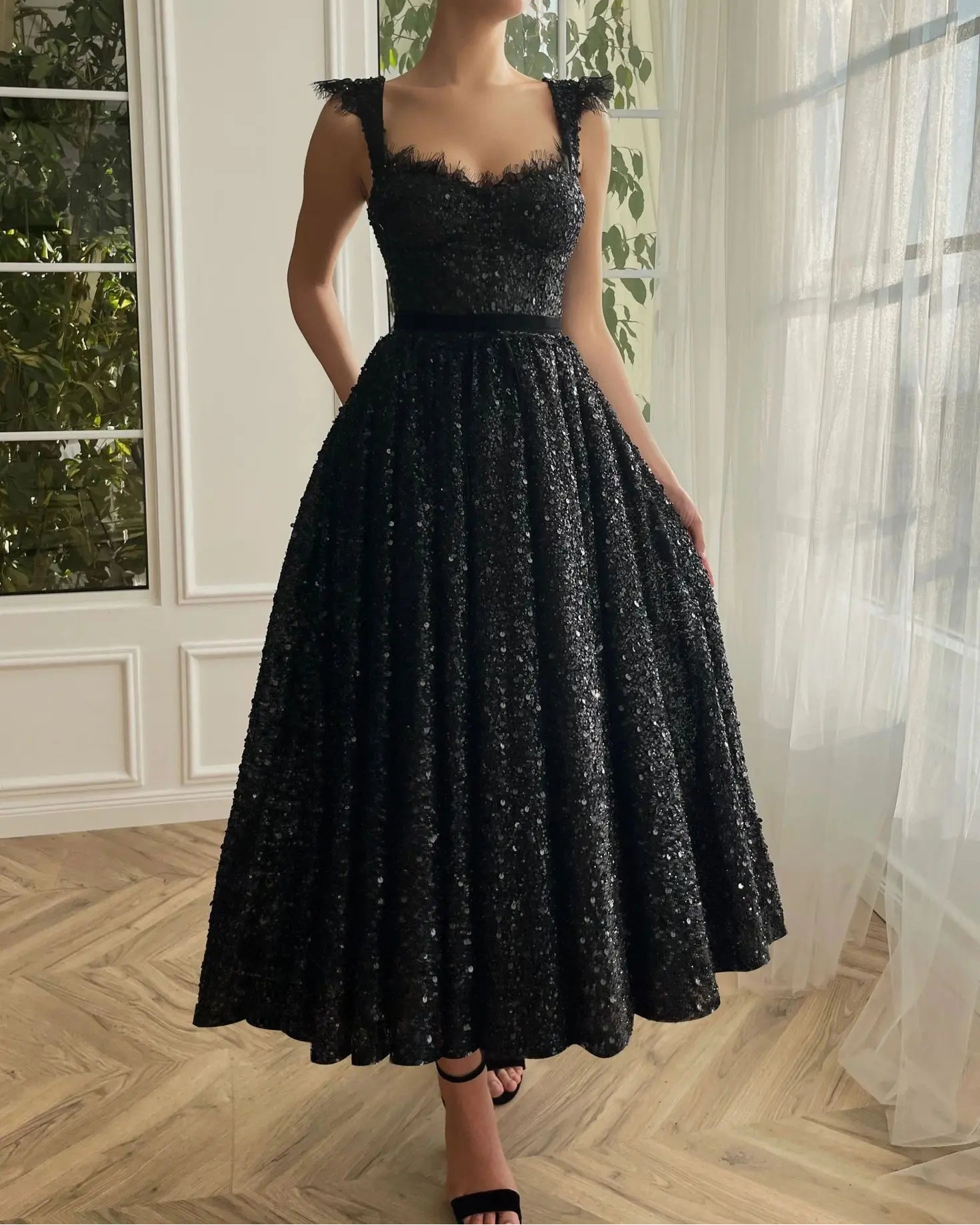A woman wearing a black sequin party dress with a v-neckline and open back, standing by a window.