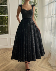 A woman wearing a black sequin party dress with a v-neckline and open back, standing by a window.