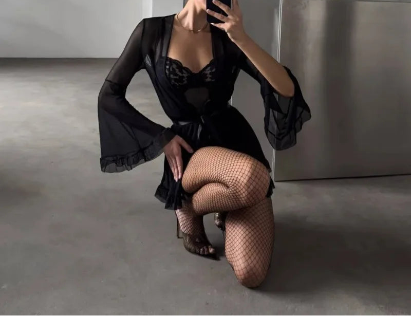 Person in a black outfit with sheer sleeves and fishnet stockings taking a mirror selfie.