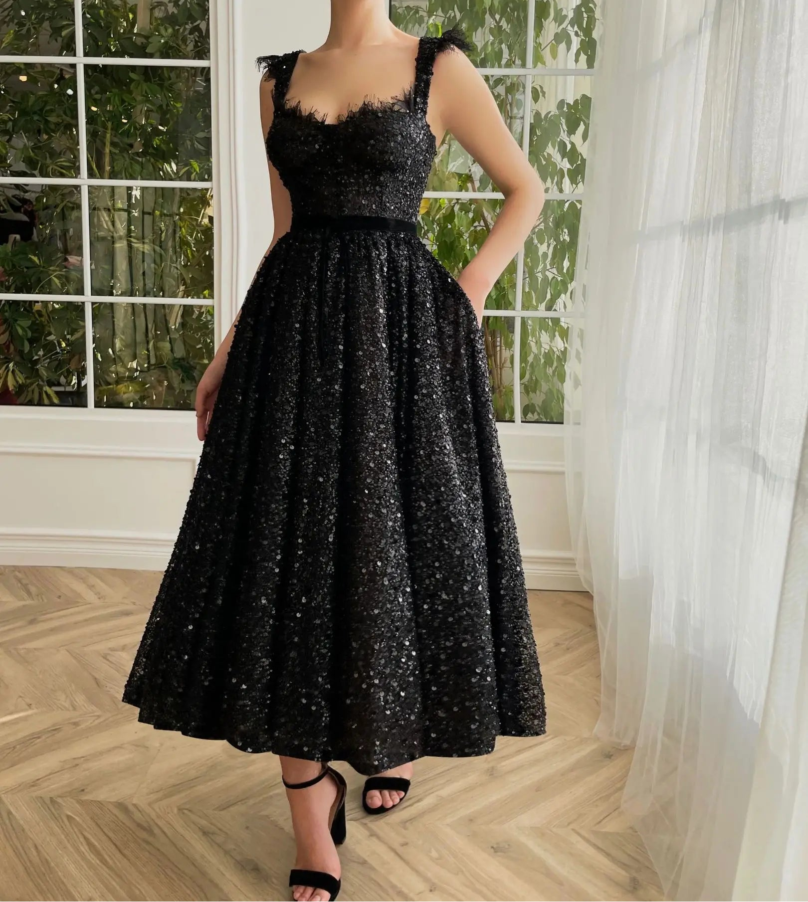A woman wearing a black sequin party dress with a v-neckline and open back, standing by a window.