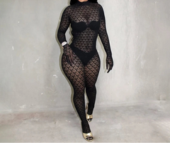A woman modeling a black sheer long-sleeve bodysuit with an allover fishnet pattern, paired with matching black gloves and gold high heels, standing against a textured gray wall.