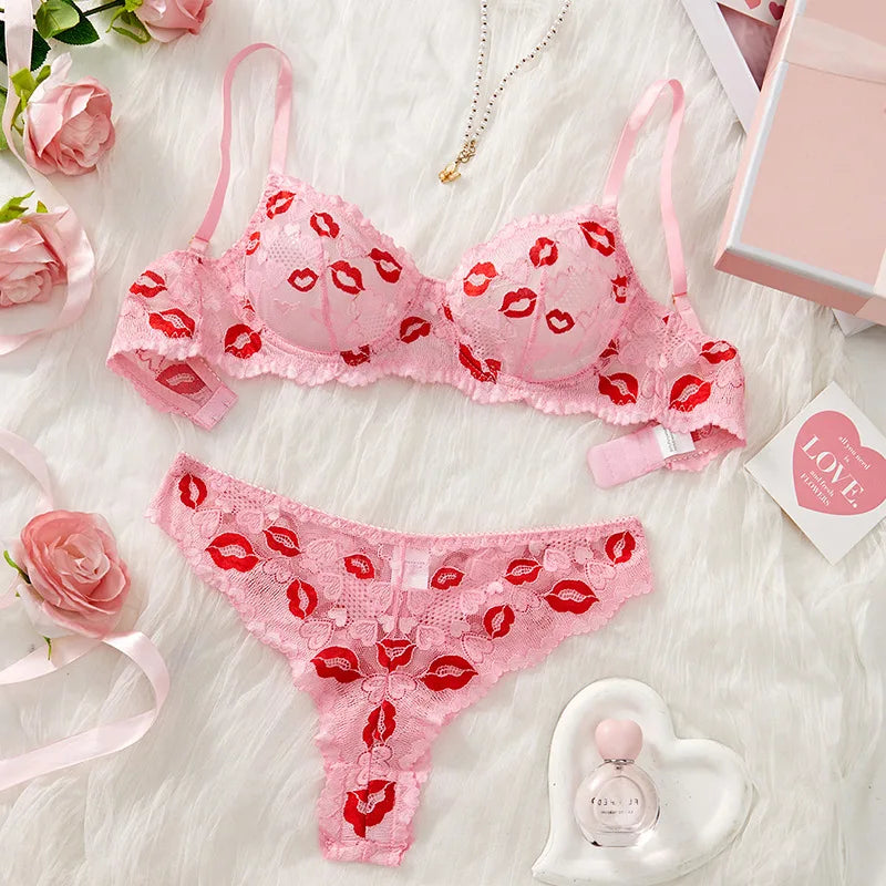 Pink lingerie set with red lip prints on a white surface with flowers and a perfume bottle.
