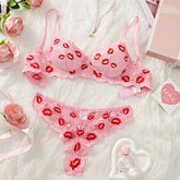 Pink lingerie set with red lip prints on a white surface with flowers and a perfume bottle.