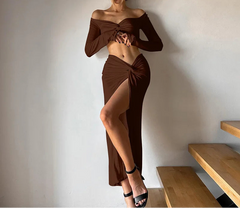 Woman in a brown off-shoulder top and skirt standing against a plain wall.