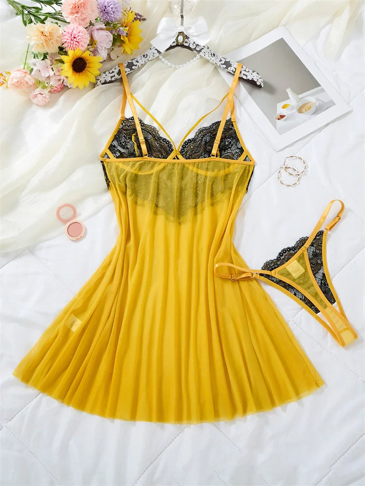 A yellow and black lace lingerie set laid flat on a white fabric background, including a sheer pleated chemise dress and matching thong, styled with flowers, a mirror, and small accessories.