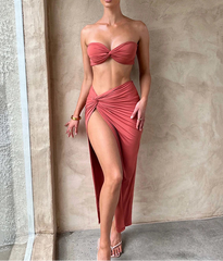 Woman in a red strapless top and high-slit skirt standing against a neutral wall.