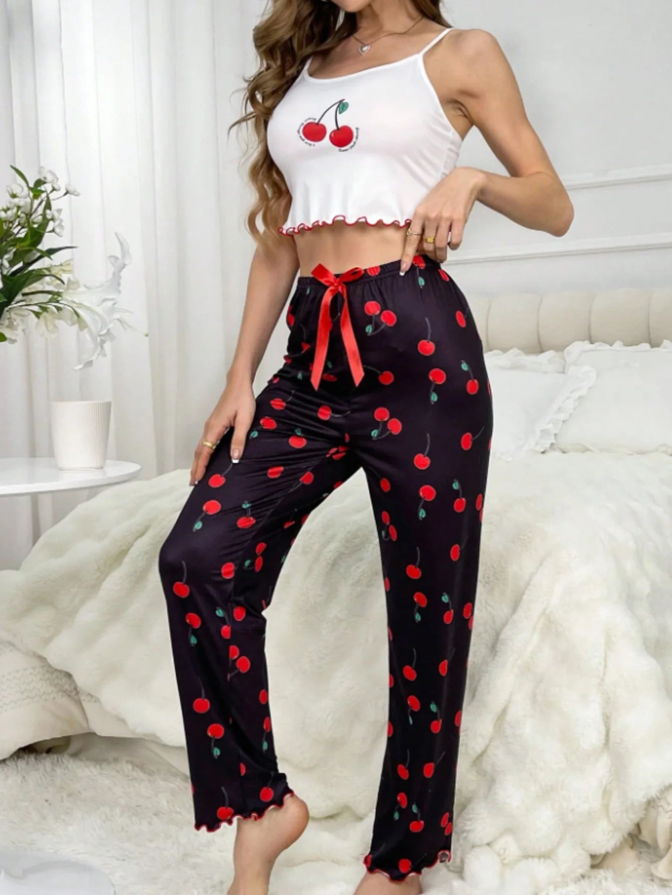 Woman wearing a white crop top with cherry design and black pants with red cherry pattern in a cozy room.