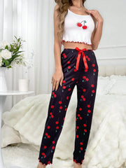 Woman wearing a white crop top with cherry design and black pants with red cherry pattern in a bedroom setting.