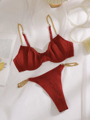 A two-piece red bikini set with gold chain straps, laid flat on a white fabric background with decorative props including a pearl necklace, a book page, and a yellow lemon slice.