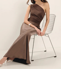 Woman in a brown one-shoulder dress sitting on a wire chair against a white background