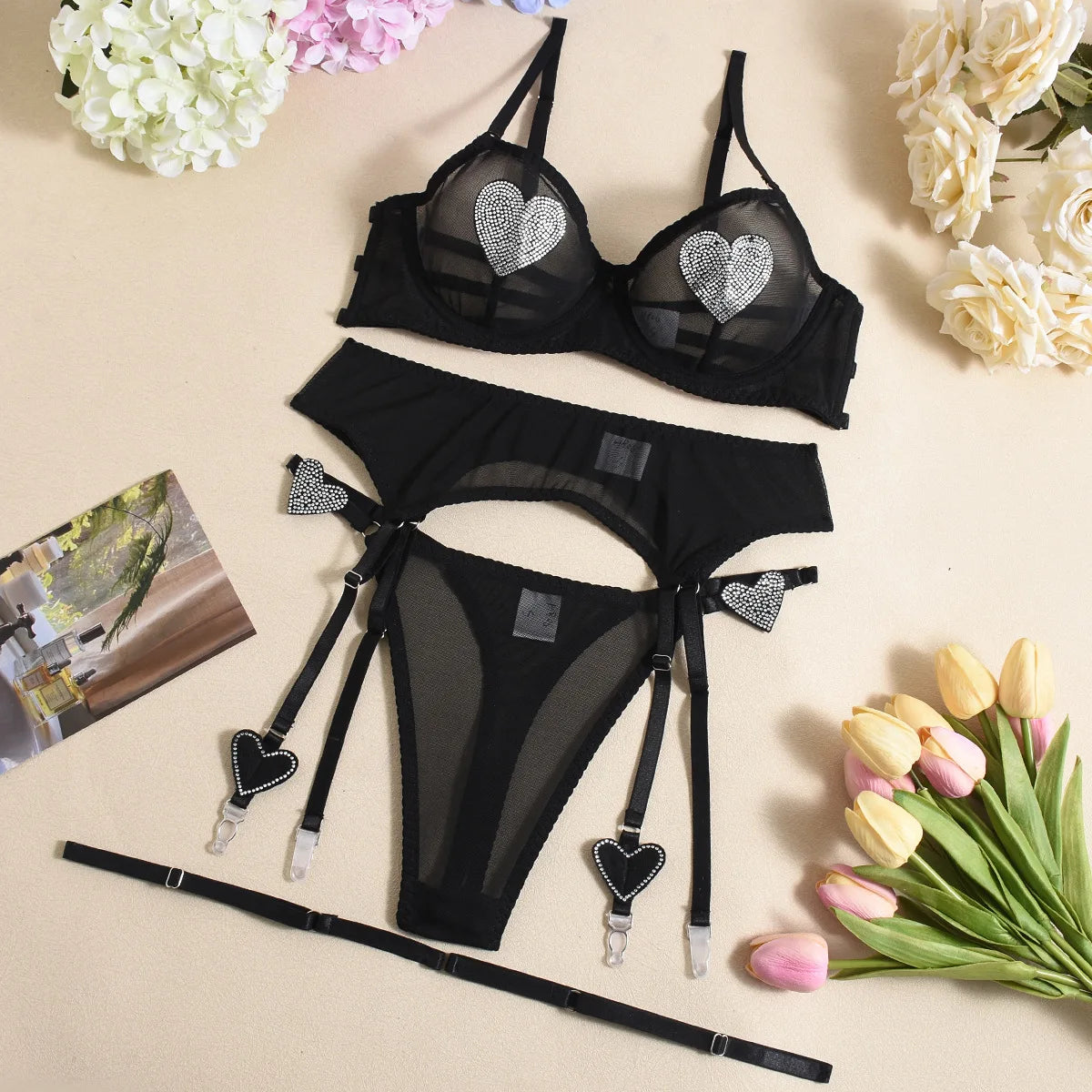 Black lingerie set with heart details on a beige surface with flowers and a magazine.