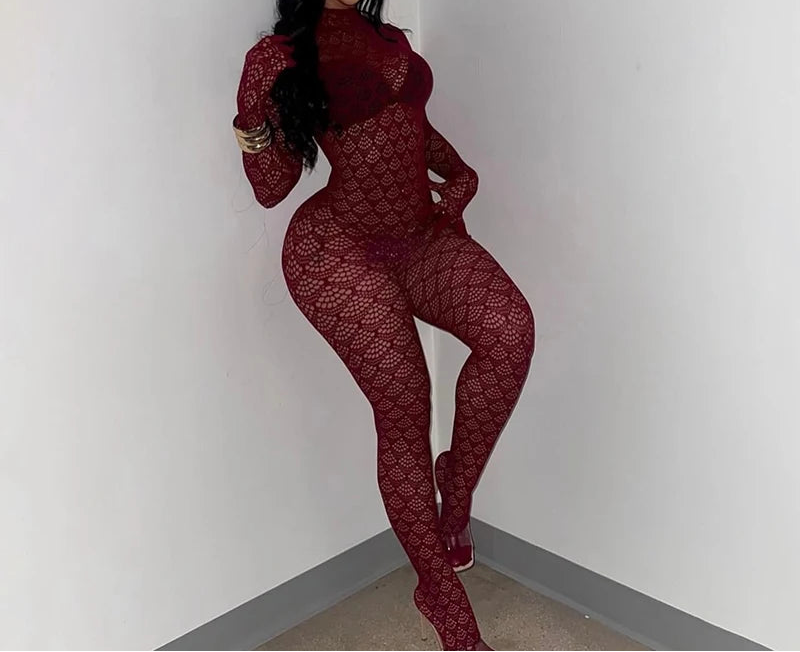 A woman wearing a long-sleeve, high-neck, full-body red fishnet jumpsuit with a textured pattern, posing in a minimalist white room corner.