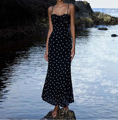 Woman in a black polka dot dress standing on a rocky beach with water and rocks in the background.