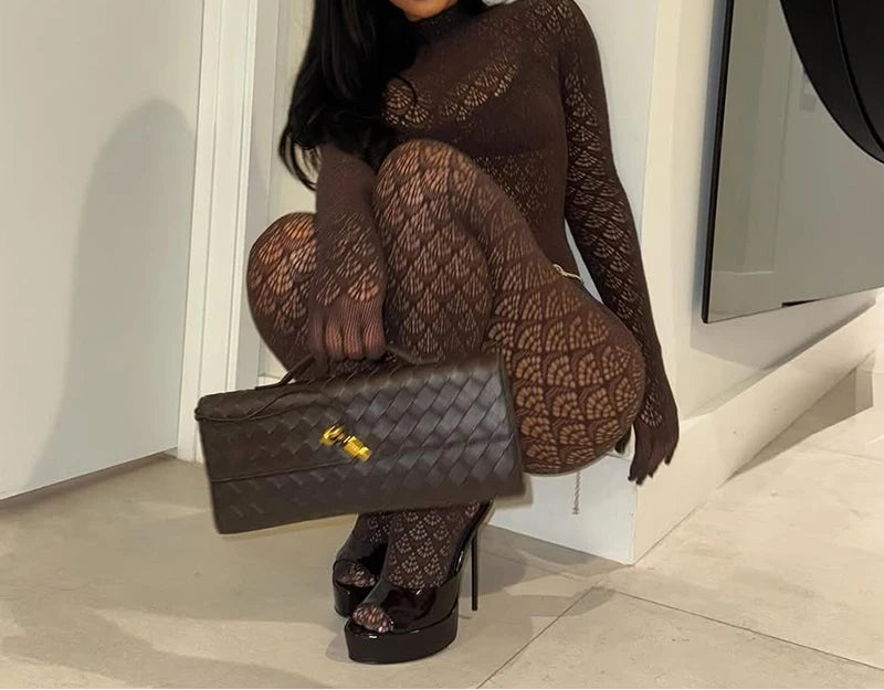 A woman wearing a dark brown patterned long-sleeve bodysuit and matching fishnet tights, posing with a woven black handbag and black platform heels in a minimalist indoor setting.