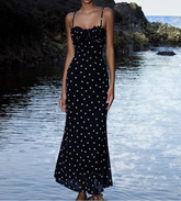 Woman in a black polka dot dress standing by a body of water with rocks.