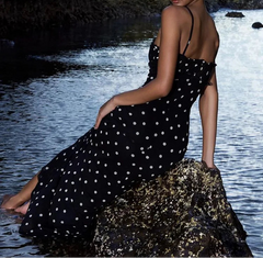 Woman in a black polka dot dress sitting on rocks by a body of water.