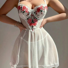 A woman wearing a white sheer babydoll lingerie chemise with red and pink rose embroidery on the bust and lace trim, posed against a neutral gray background.