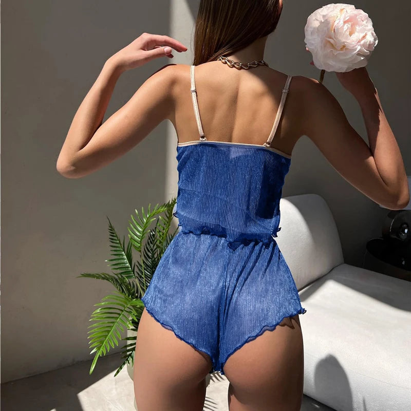 Woman wearing a blue denim romper holding a white flower, standing in a room with a plant and chair.