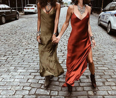 Two women walking down a street wearing sleeveless, V-neck maxi dresses in different colors, with one dress in green and the other in red, both with side pockets and adjustable straps.