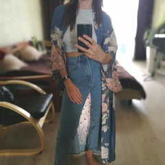 A person wearing a long kimono with floral embroidery, paired with a white tank top and blue jeans, taking a mirror selfie.