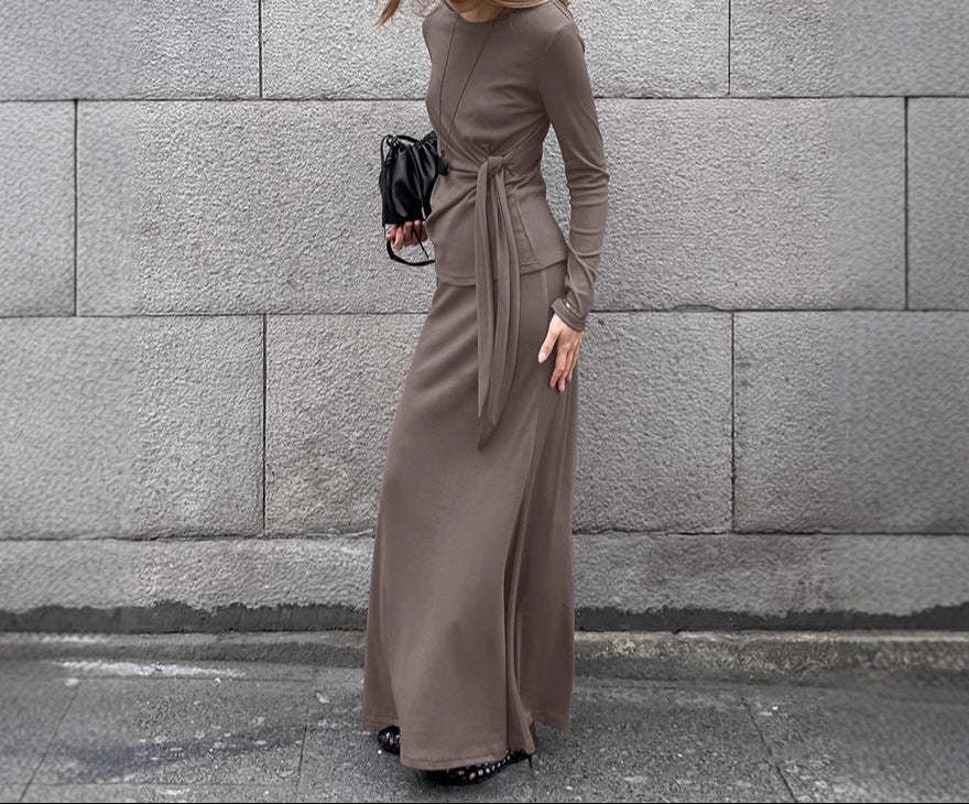 Woman in a long brown dress standing against a gray stone wall.