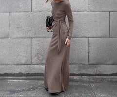 Woman in a long brown dress standing against a gray stone wall.
