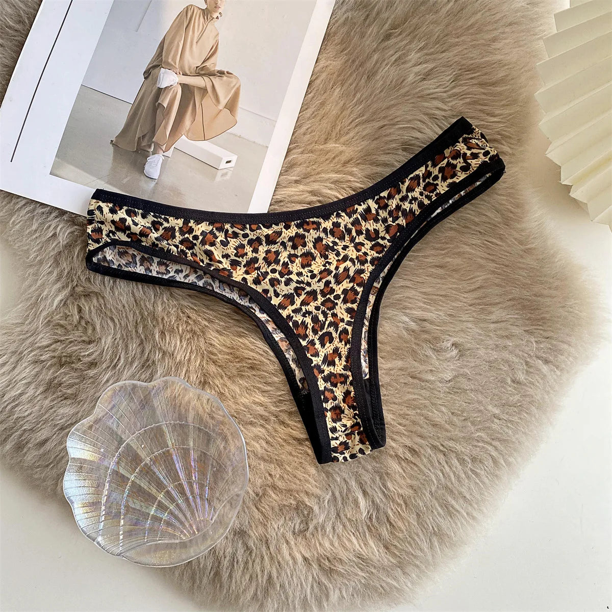 Leopard-print G-string with black trim laid flat on a textured beige surface beside a magazine and shell.