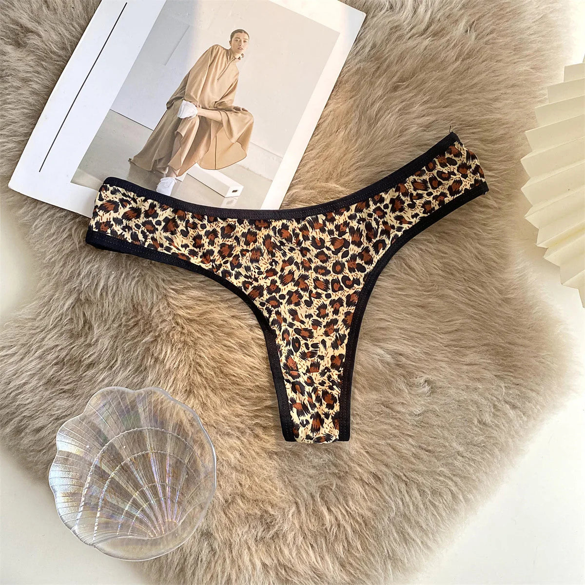 Leopard-print G-string with black trim displayed flat on a textured beige surface beside a magazine and shell.