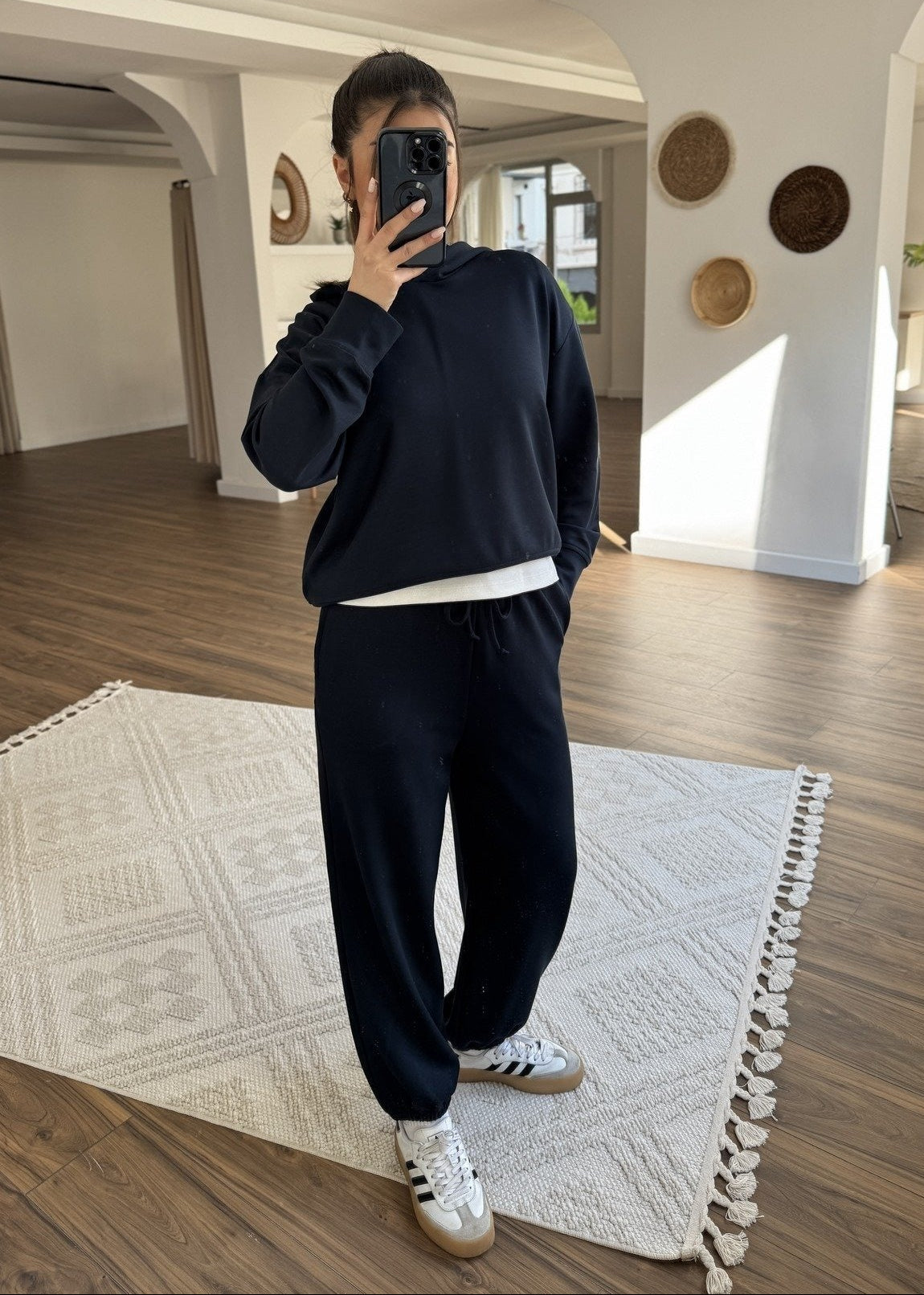 Person taking a mirror selfie wearing a black tracksuit and Adidas sneakers in a room with wooden flooring and white walls.