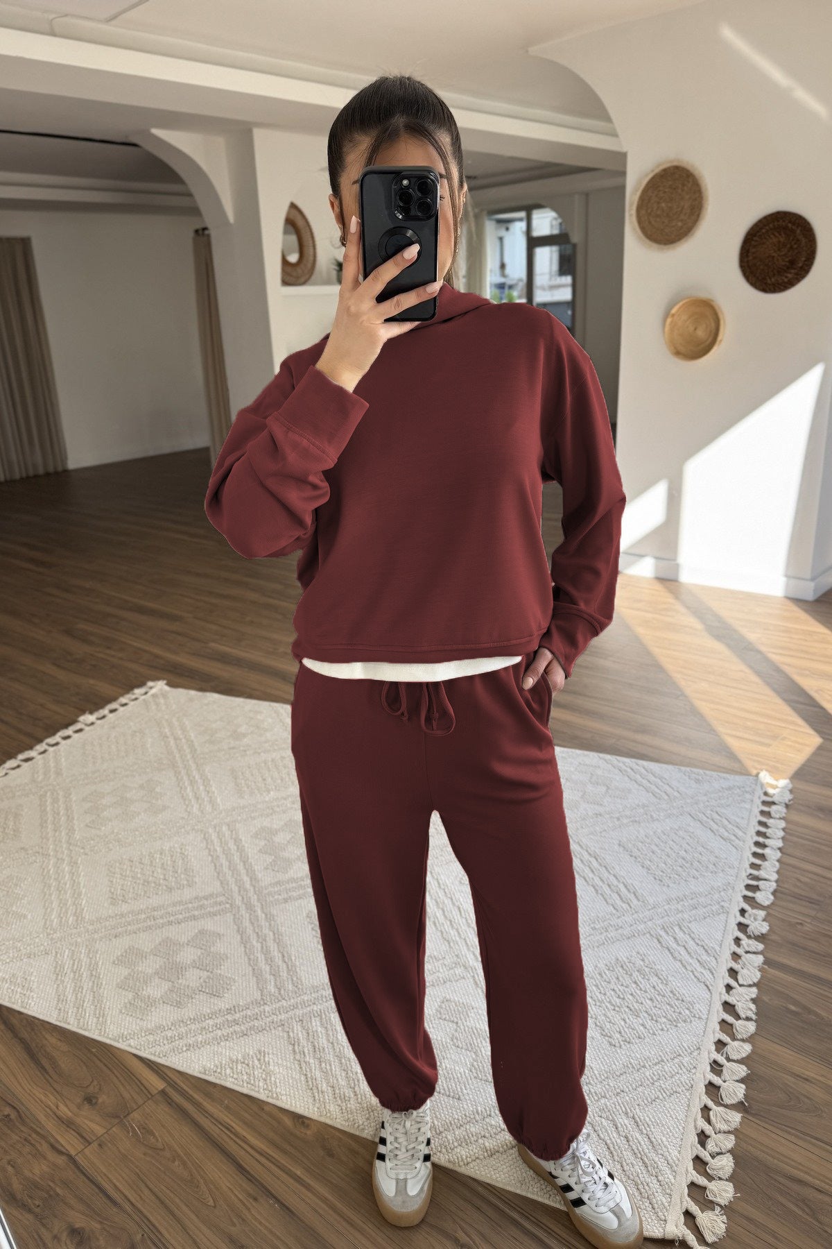 Person wearing a burgundy tracksuit taking a mirror selfie in a room with wooden flooring and decorative wall elements.