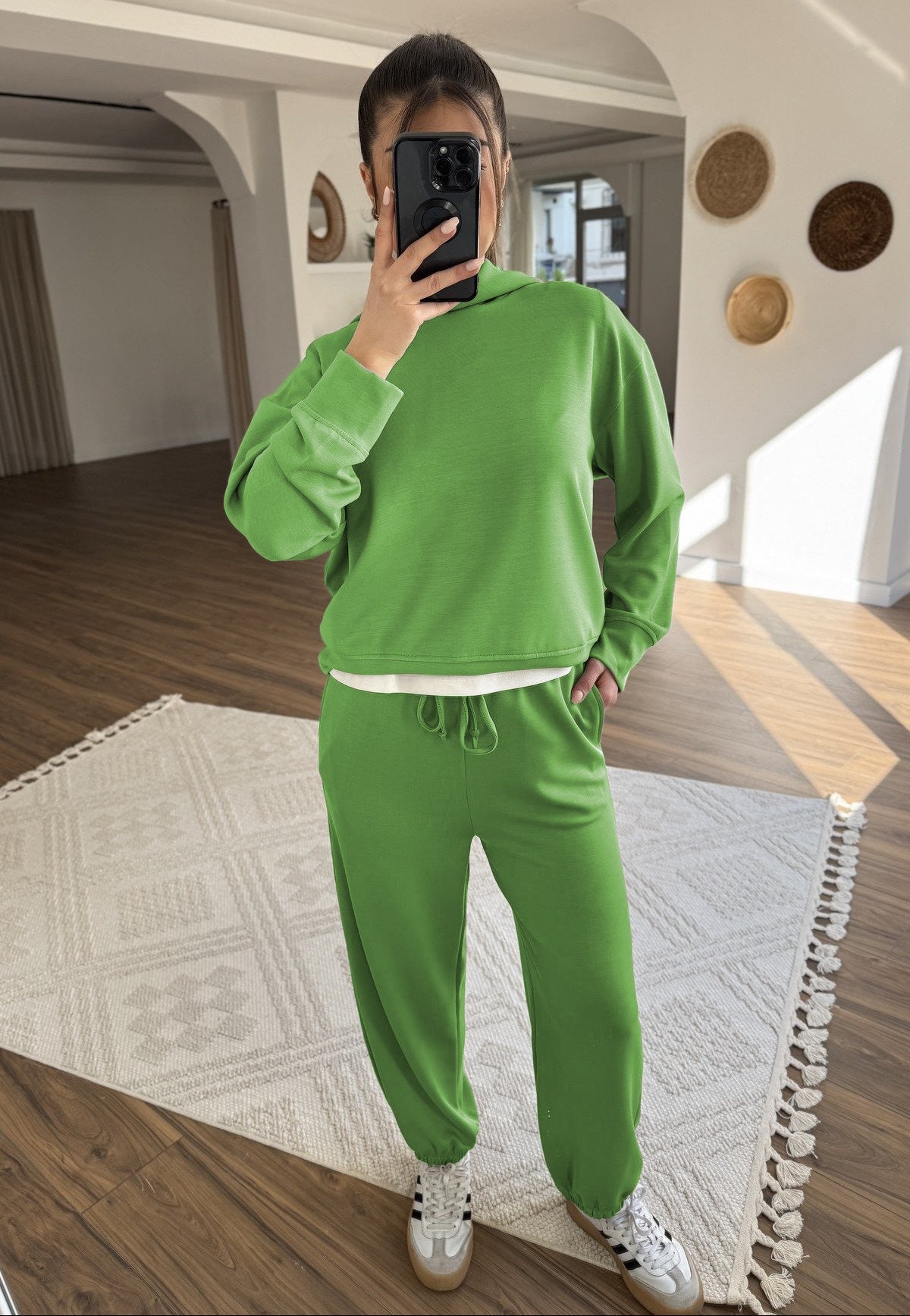Person wearing a green tracksuit taking a mirror selfie in a room with wooden floor and white walls.