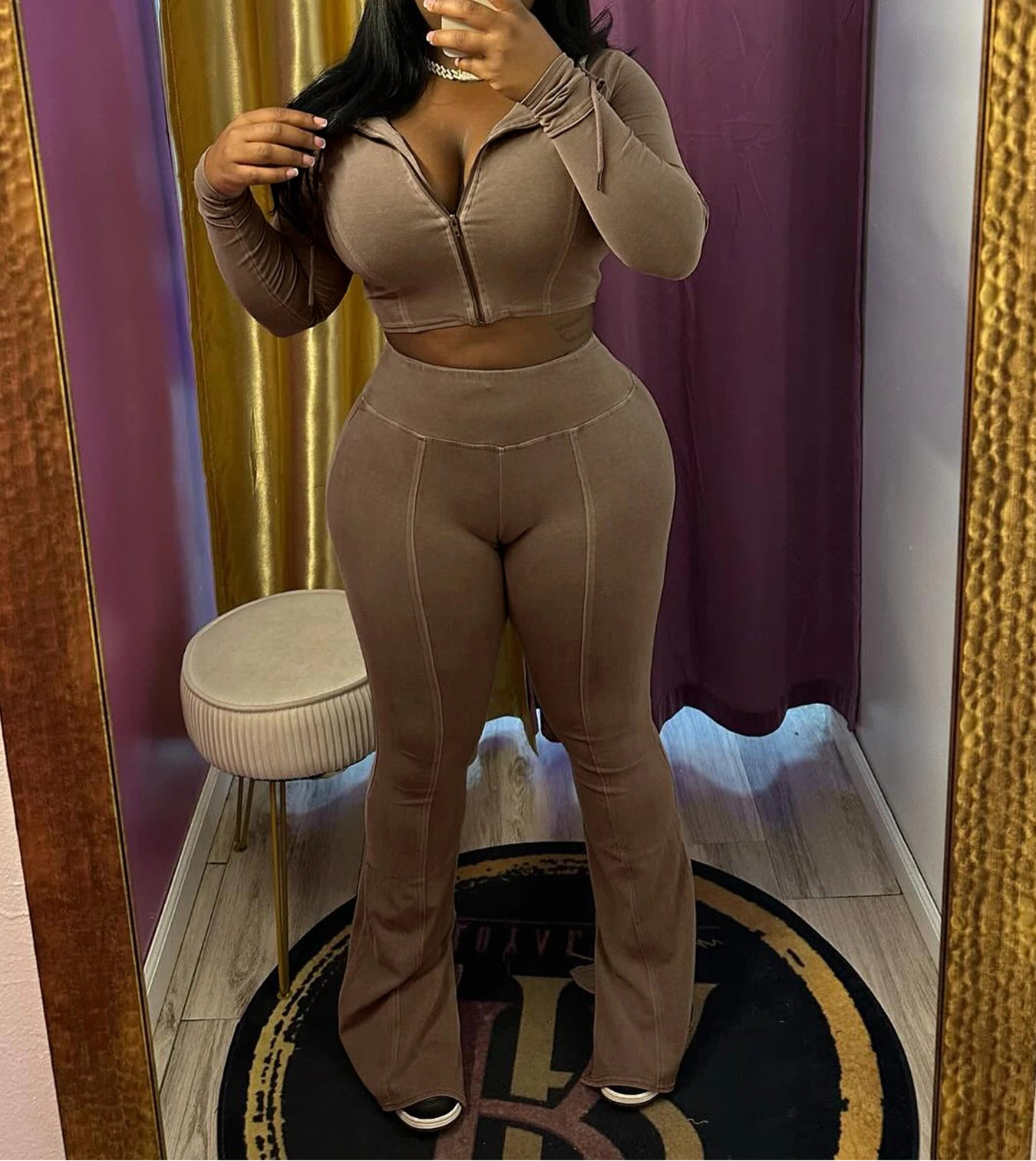 Person wearing a beige outfit taking a mirror selfie in a room with gold curtains and a small stool.