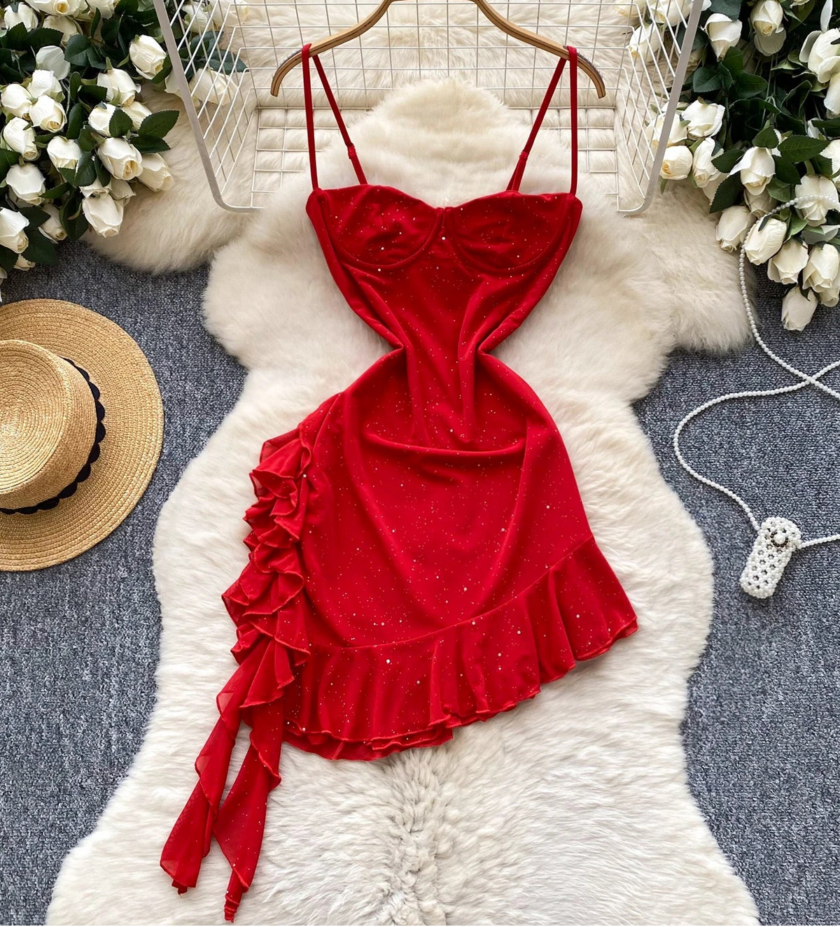 Red dress with ruffles on a white fur rug surrounded by flowers and a hat.