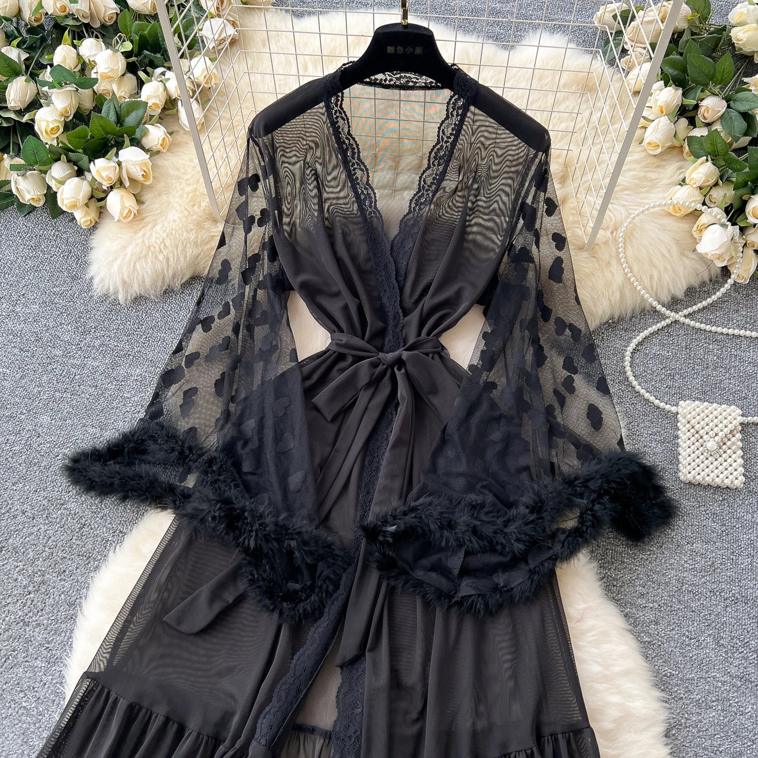 Black lace lingerie set with feathered sleeves on a hanger, surrounded by decorative flowers and fur.