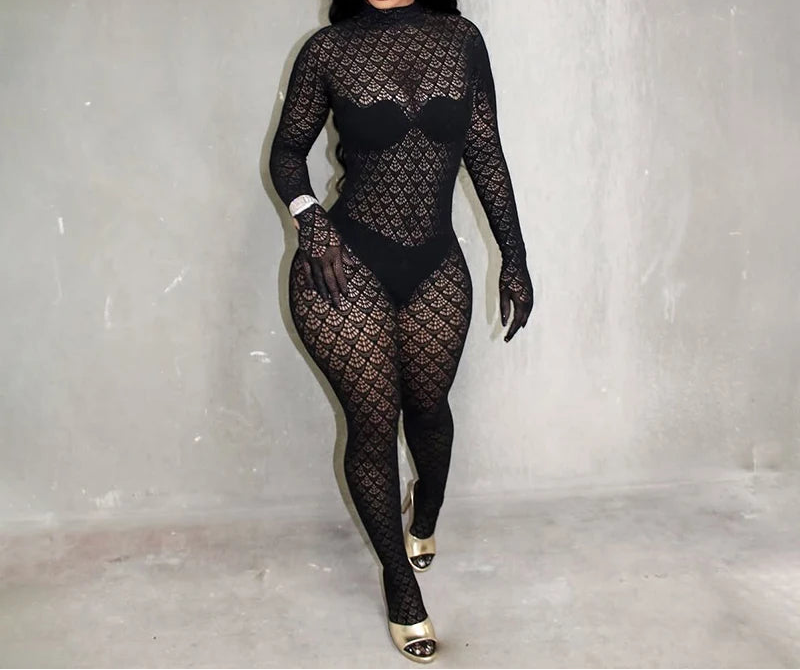 A woman modeling a black sheer long-sleeve bodysuit with an allover fishnet pattern, paired with matching black gloves and gold high heels, standing against a textured gray wall.
