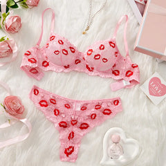 Pink lingerie set with red lip prints on a white surface with flowers and a perfume bottle.