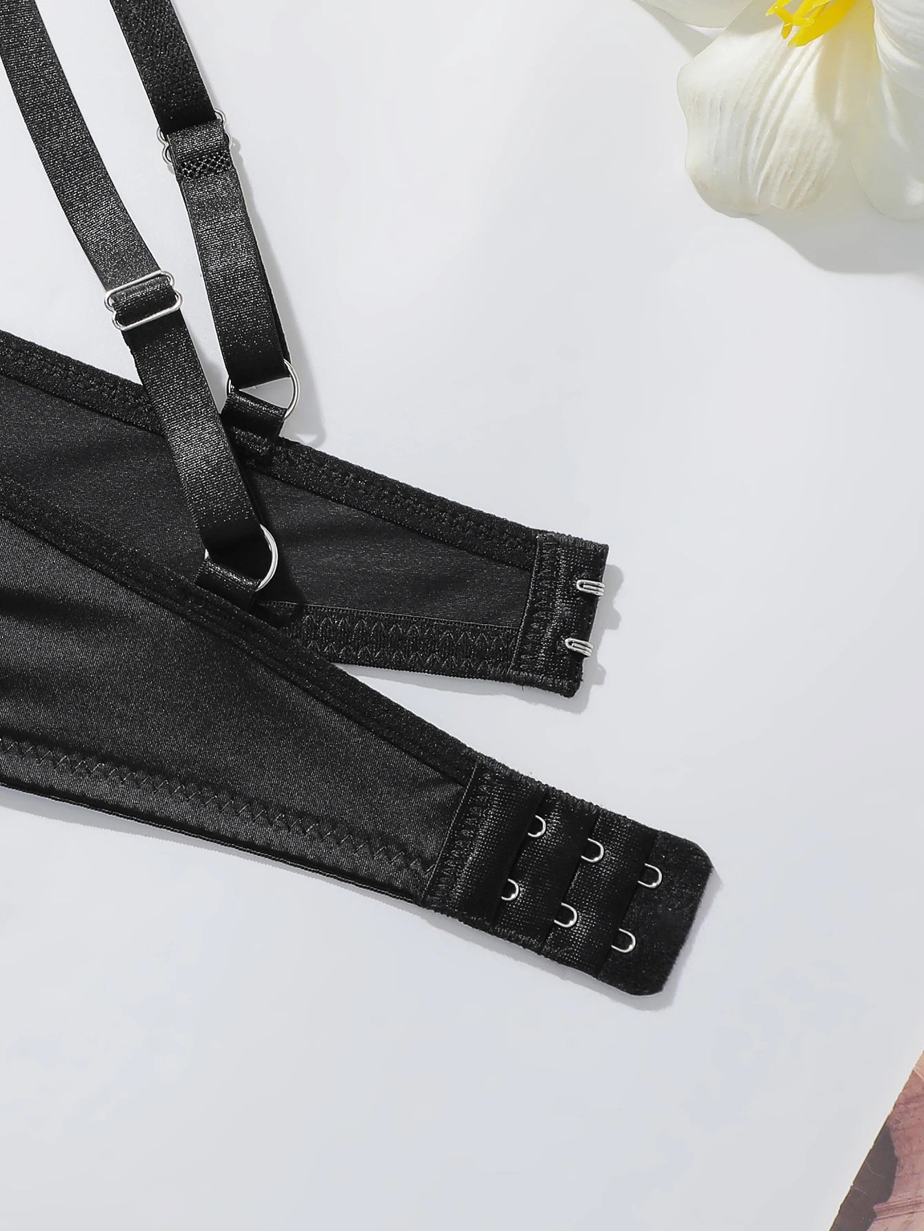 Black bra strap with adjustable hook-and-eye closure and metal hardware, photographed on a clean white background with a small white flower prop in the corner.