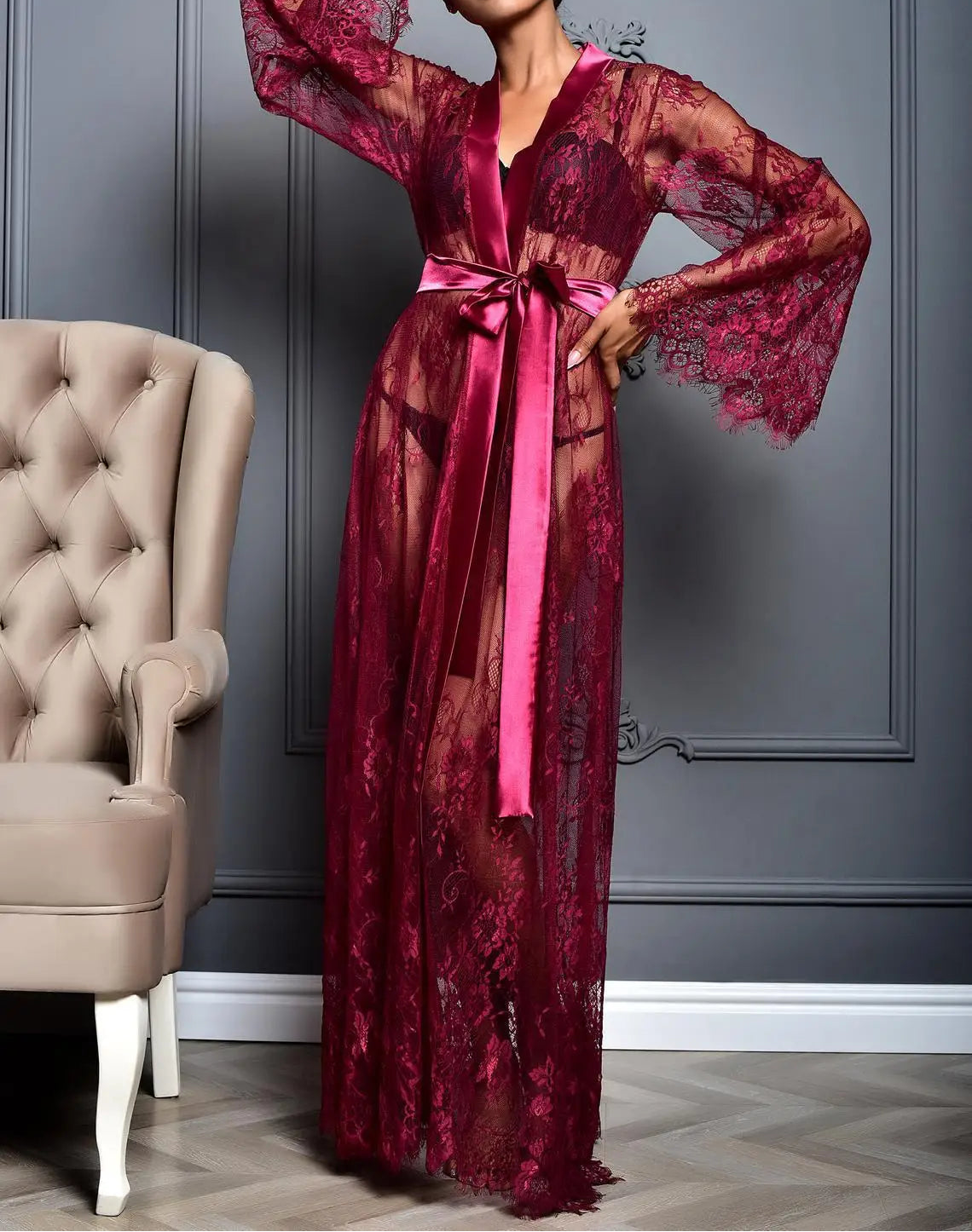 Woman wearing a burgundy lace dress with a belt in a room with gray walls and a beige chair.