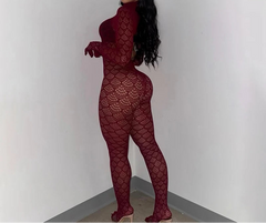 A woman modeling a long-sleeve, high-neck, full-body lace jumpsuit in a deep burgundy red, standing in a plain white room corner.