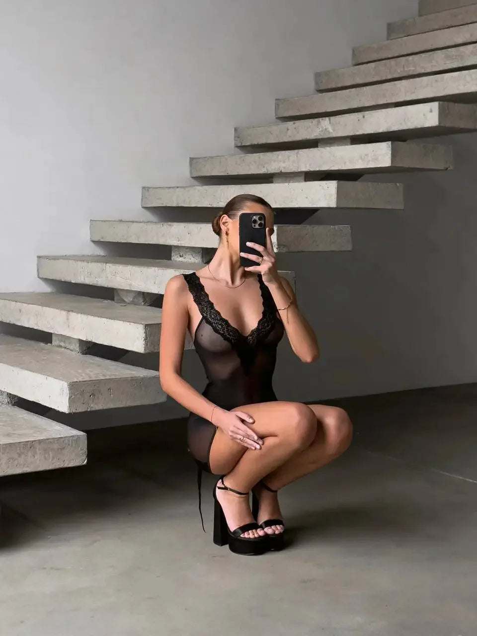 Person in black lingerie taking a mirror selfie against a modern staircase.