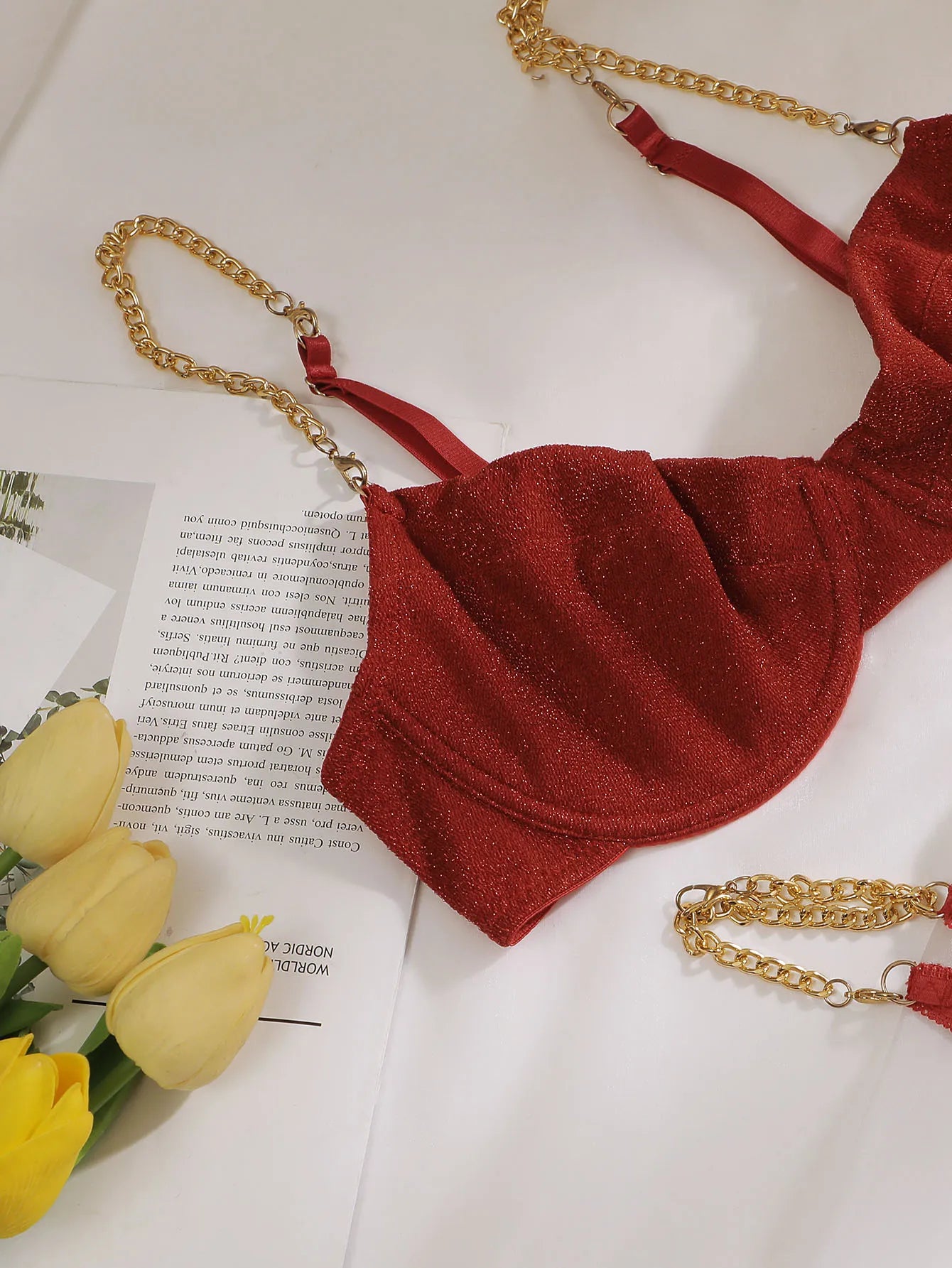 A red velvet bralette with gold chain straps, styled on a white surface with yellow tulips and an open book.