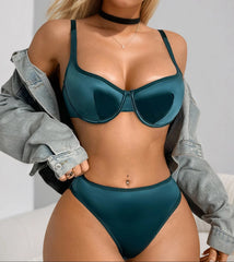 A woman modeling a two-piece satin lingerie set in a deep teal green color, featuring a bra and matching high-cut bottoms, styled with an off-the-shoulder light-wash denim jacket.