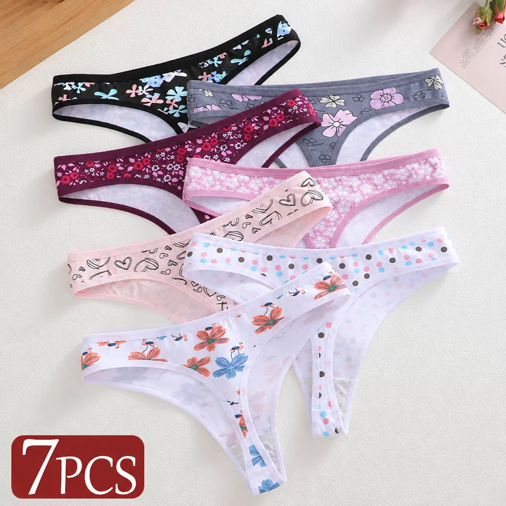 Seven pairs of women's sheer lace thong panties arranged diagonally on a light beige background. The set includes various colors and floral patterns: black with blue flowers, gray with pink flowers, burgundy with red flowers, light pink with white flowers, light pink with black line art, white with orange and blue flowers, and white with blue and pink polka dots.