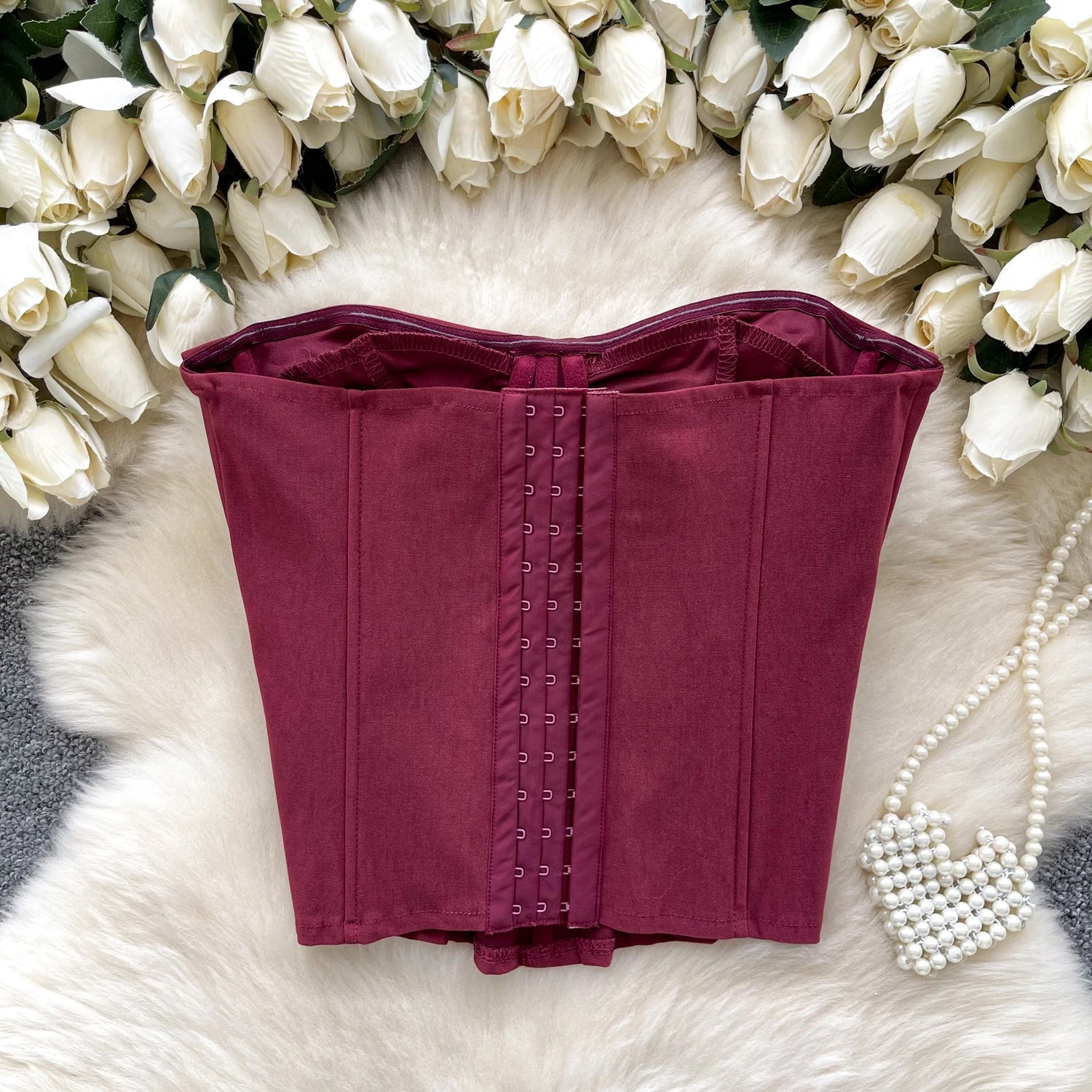 Maroon corset on a white fur surface with white roses and pearls in the background