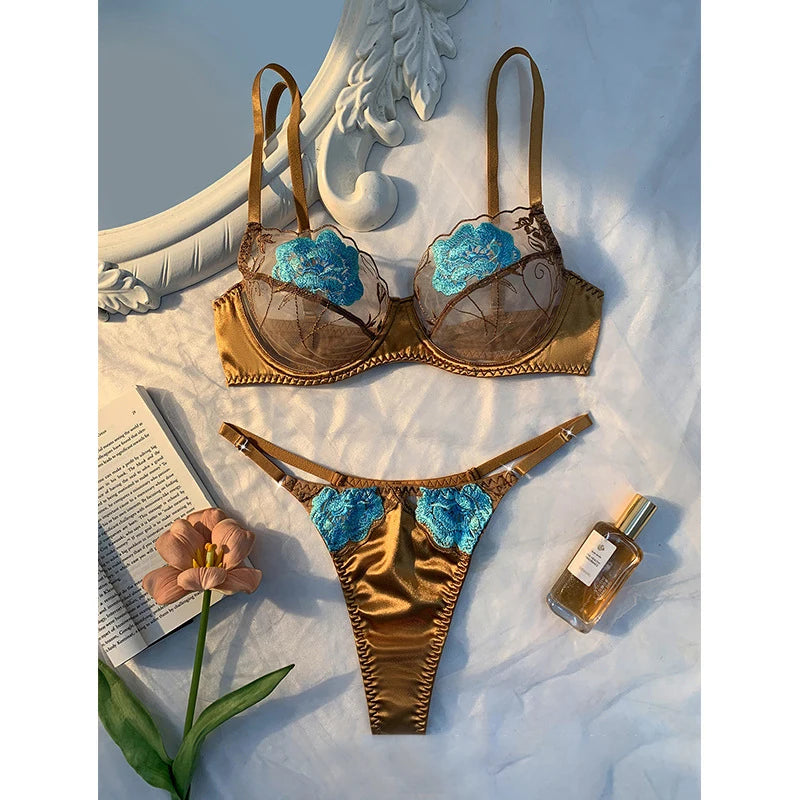 Gold bra set with blue floral details on a white surface with a book and a flower.