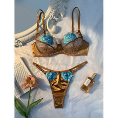 Gold bra set with blue floral details on a white surface with a book and a flower.