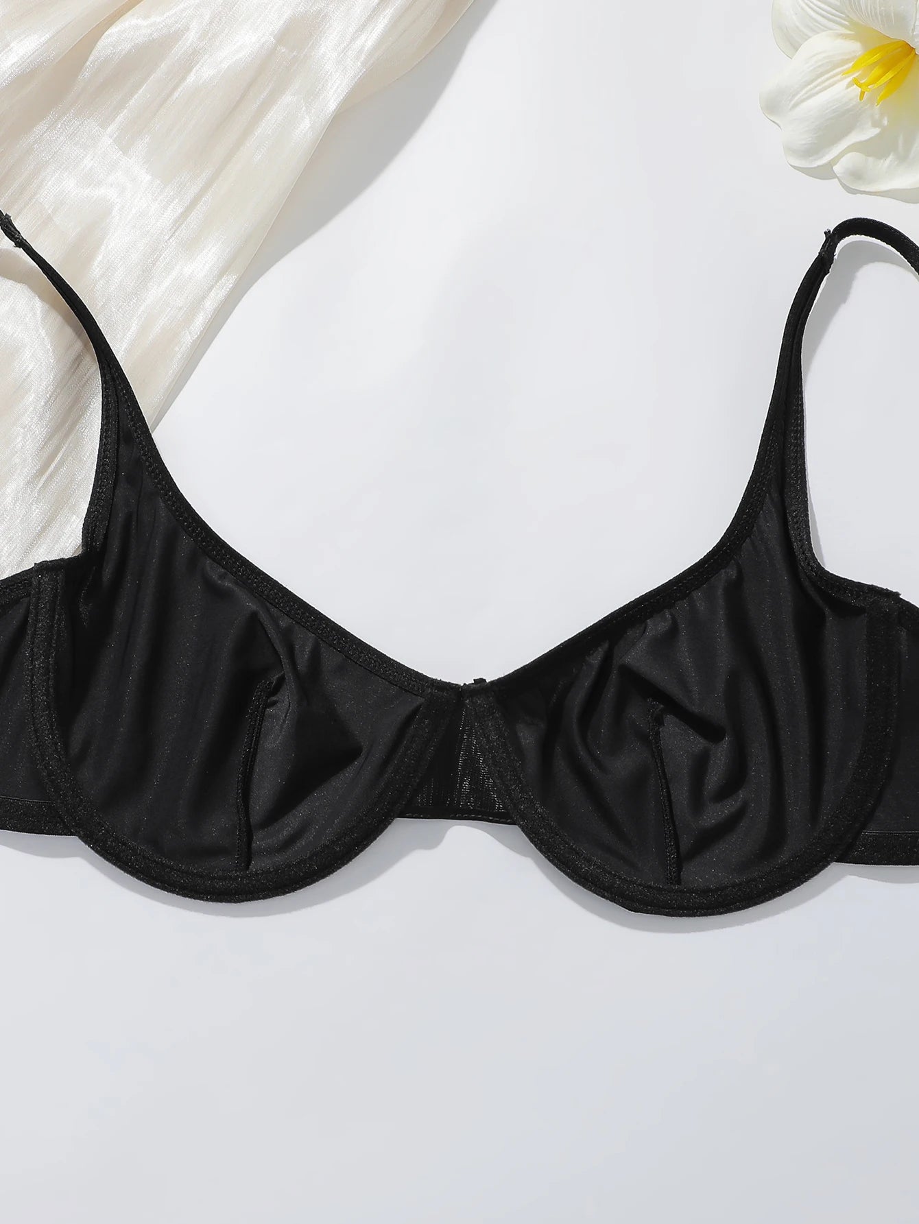 Black underwire bra with thin adjustable shoulder straps, laid flat on a white background with decorative white fabric and a white flower in the corners.