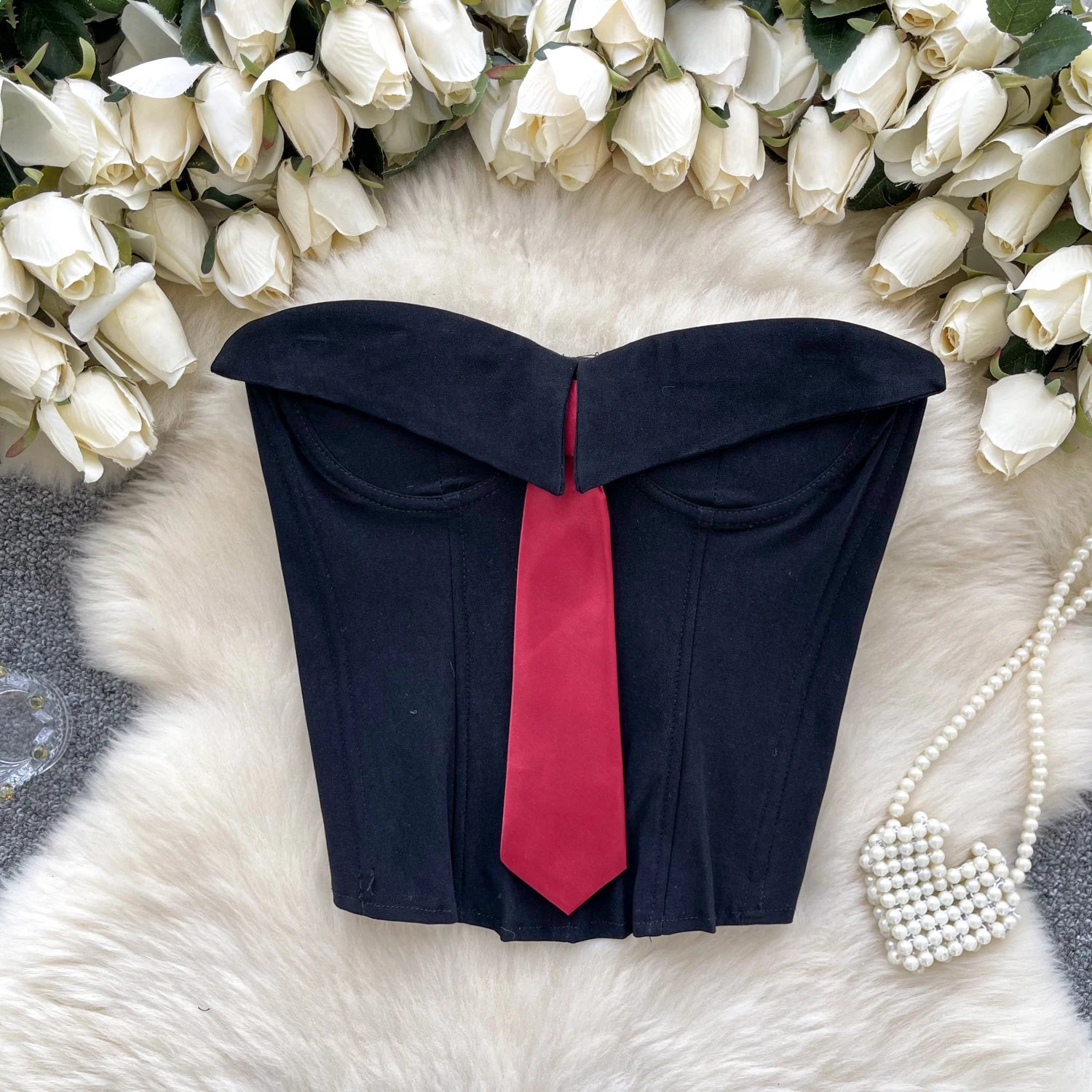 Black bustier with a red tie on a white fur surface surrounded by white flowers.